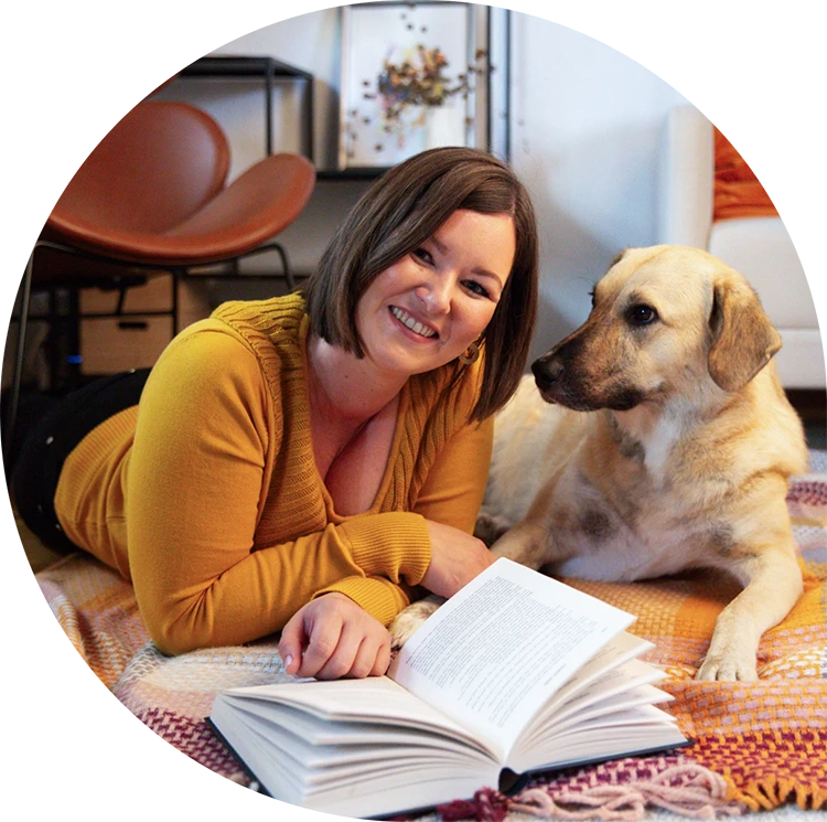 Birgit Spalt-Zoidl liegt neben ihrem Hund auf einer bunten Decke und blickt lächelnd in die Kamera, während vor ihr ein Buch aufgeschlagen ist.